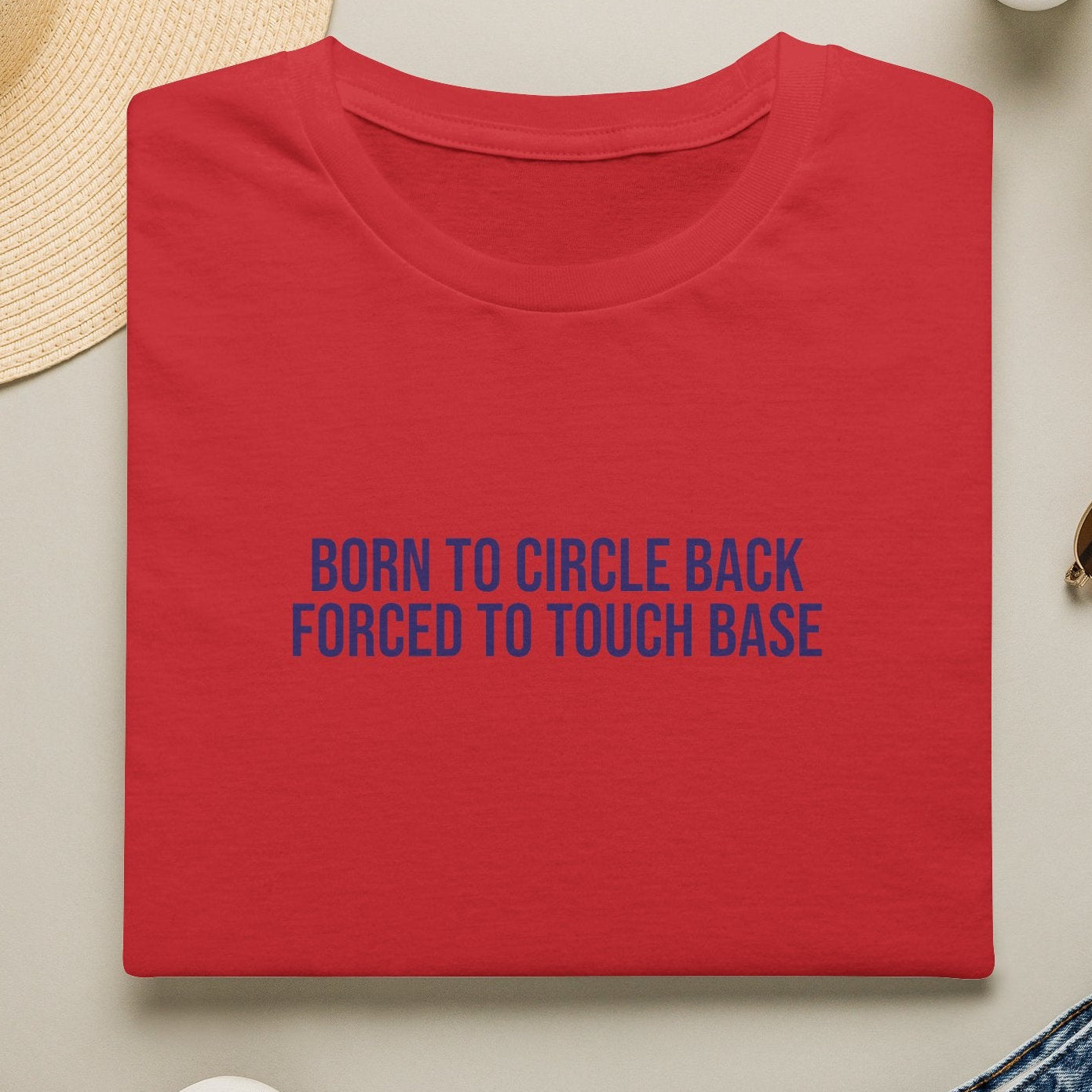 Red t-shirt with blue text 'Born to Circle Back Forced to Touch Base' on a light surface with casual items.