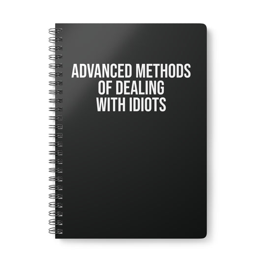 Black notebook with white text 'Advanced Methods of Dealing with Idiots' on a white background