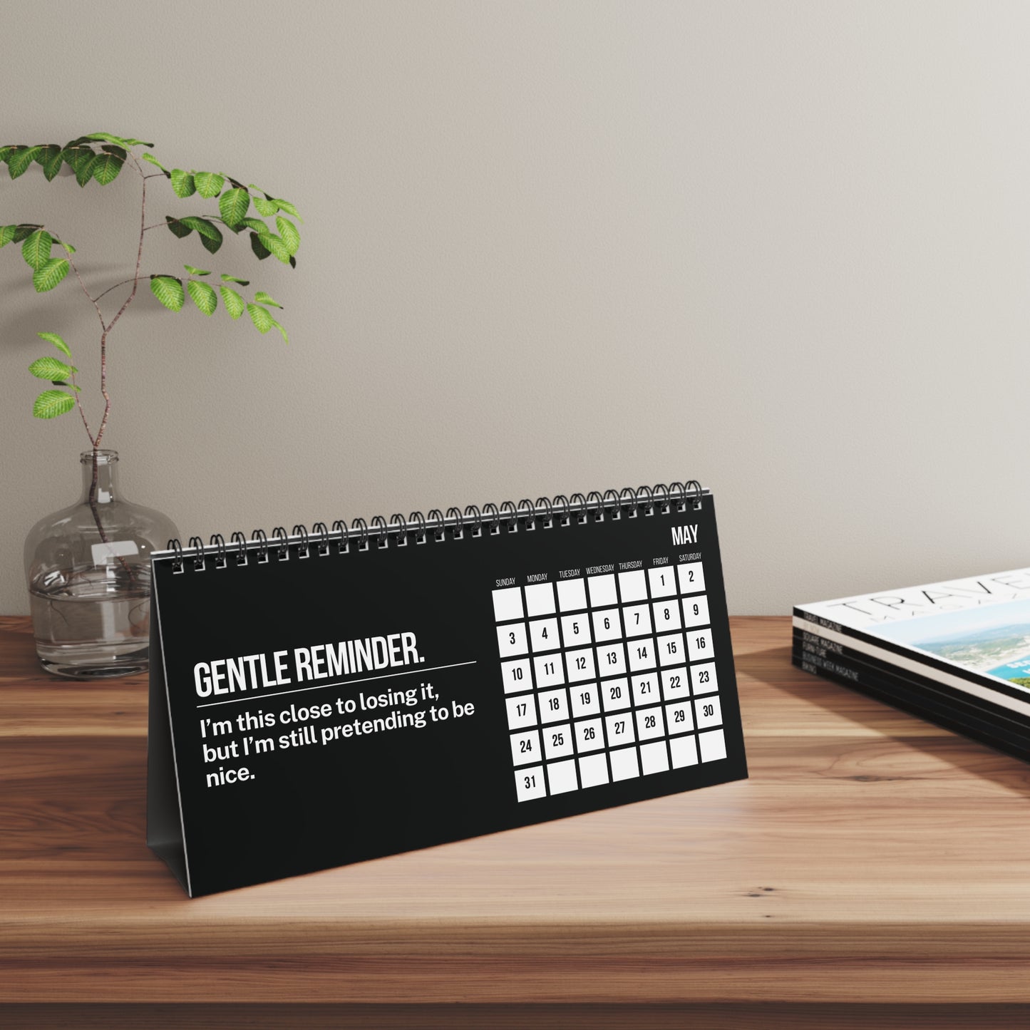 Desk calendar with humorous message on a wooden surface with a plant and magazine in the background.