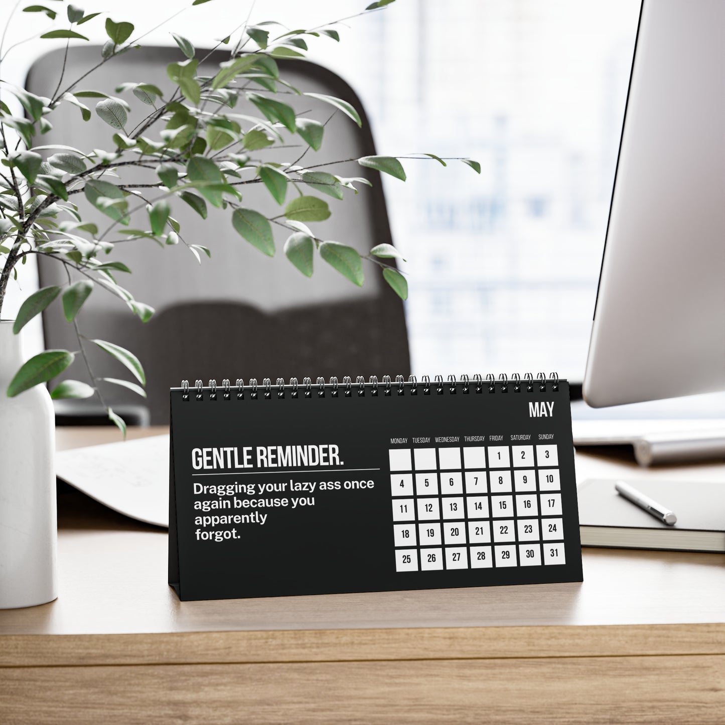 Desk calendar with humorous message on a desk with office items and plant.