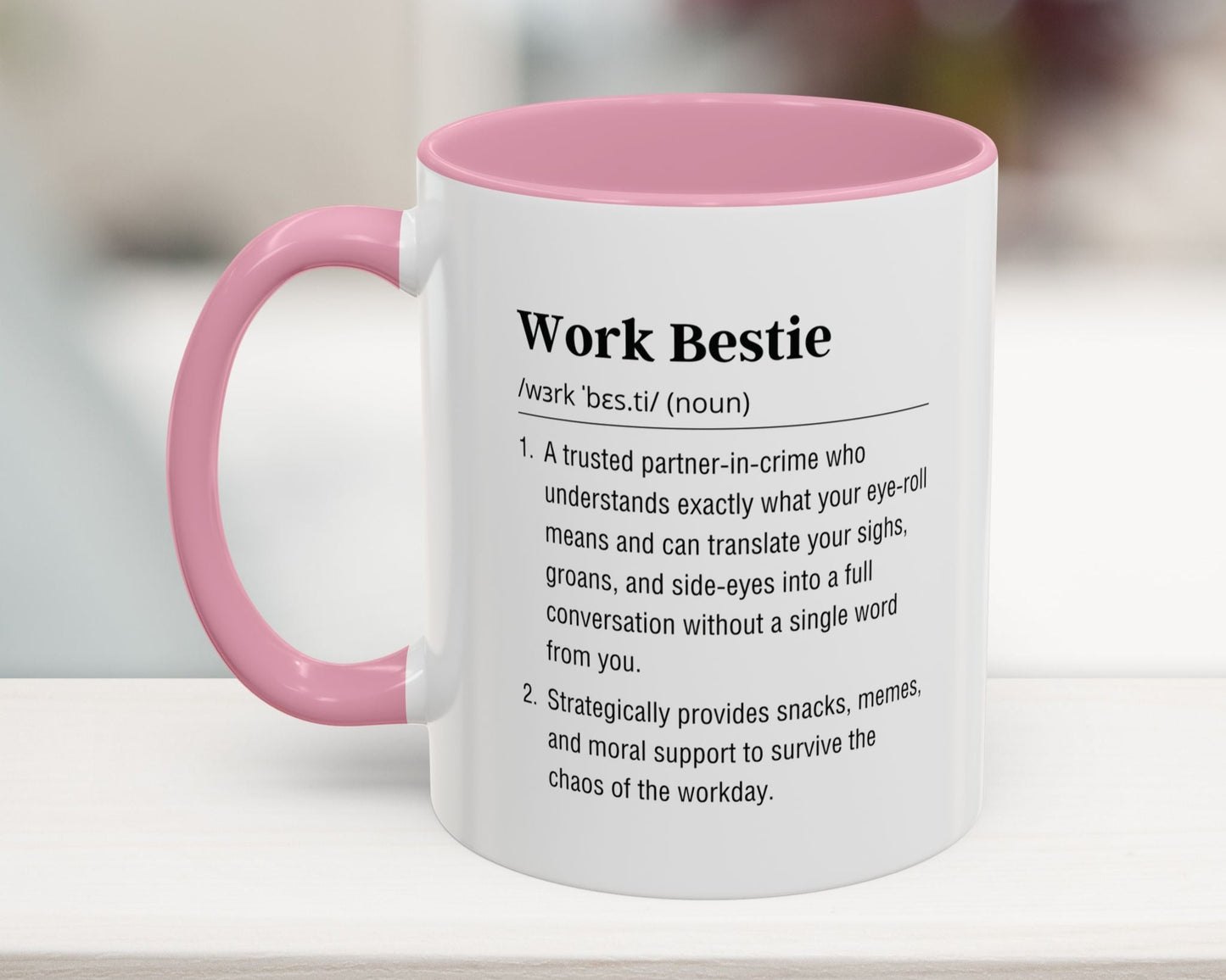 White mug with pink handle featuring text definition of 'Work Bestie' on a blurred background