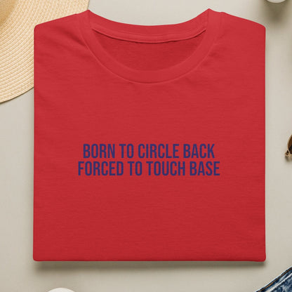 Red t-shirt with blue text 'Born to Circle Back Forced to Touch Base' on a light surface with casual items.