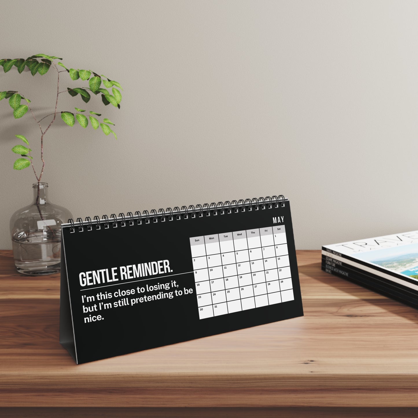 A spiral-bound desk calendar standing on a wooden table, featuring a black cover with white text that reads 'GENTLE REMINDER. I'm this close to losing it, but I'm still pretending to be nice.