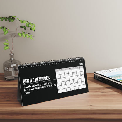 A spiral-bound desk calendar standing on a wooden table, featuring a black cover with white text that reads 'GENTLE REMINDER. I'm this close to losing it, but I'm still pretending to be nice.