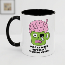A white ceramic coffee mug with a black handle and black interior, featuring a cartoon graphic of a green coffee cup with a pink brain on top and the text 'DEAD AT WORK BEFORE MY MORNING COFFEE'.