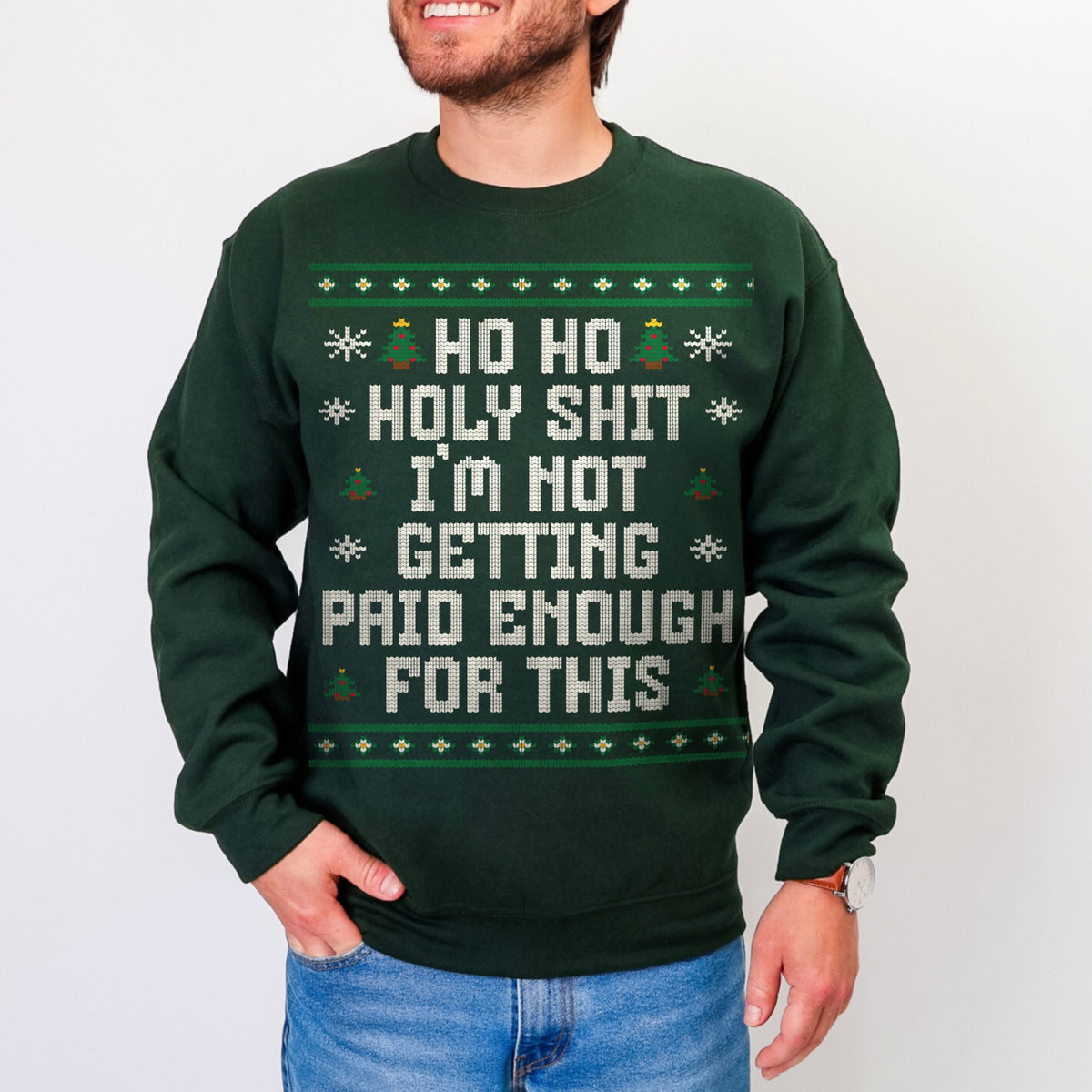A man wearing a dark green crewneck sweatshirt with a humorous Christmas-themed graphic that reads 'HO HO HOLY SHIT I'M NOT GETTING PAID ENOUGH FOR THIS' in white text, styled like a classic ugly holiday sweater.