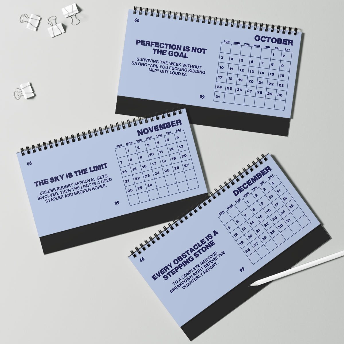Three spiral-bound monthly calendar notebooks for October, November, and December are arranged on a light gray surface with binder clips and a pen nearby. The pages are pale blue with dark navy text and grids, and the notebooks have dark gray spiral binding.