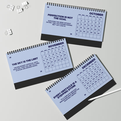 Three spiral-bound monthly calendar notebooks for October, November, and December are arranged on a light gray surface with binder clips and a pen nearby. The pages are pale blue with dark navy text and grids, and the notebooks have dark gray spiral binding.