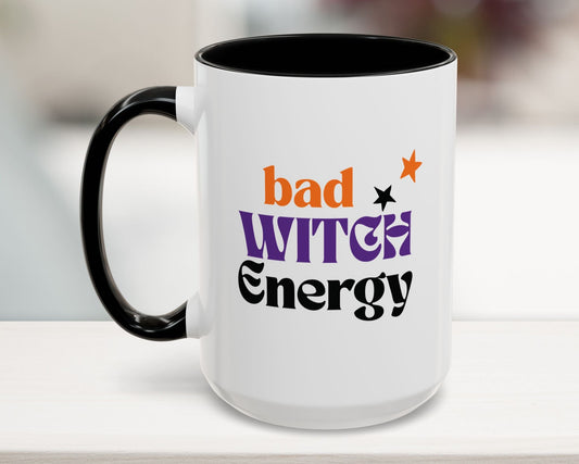 White mug with black interior and handle, featuring colorful text 'bad WITCH Energy' on a blurred background.