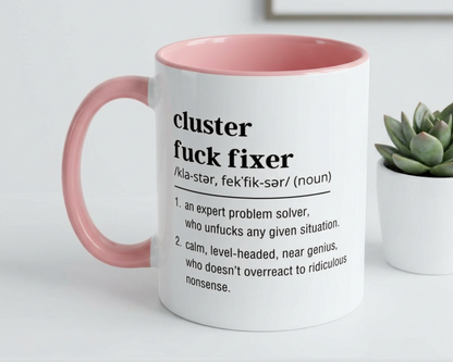 White mug with pink handle featuring a definition of 'cluster fuck fixer' on a white surface with a plant in the background.