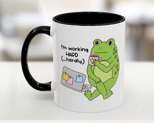 Mug with a green frog holding a cup and text 'I'm working HARD (...hardly)' on a blurred background
