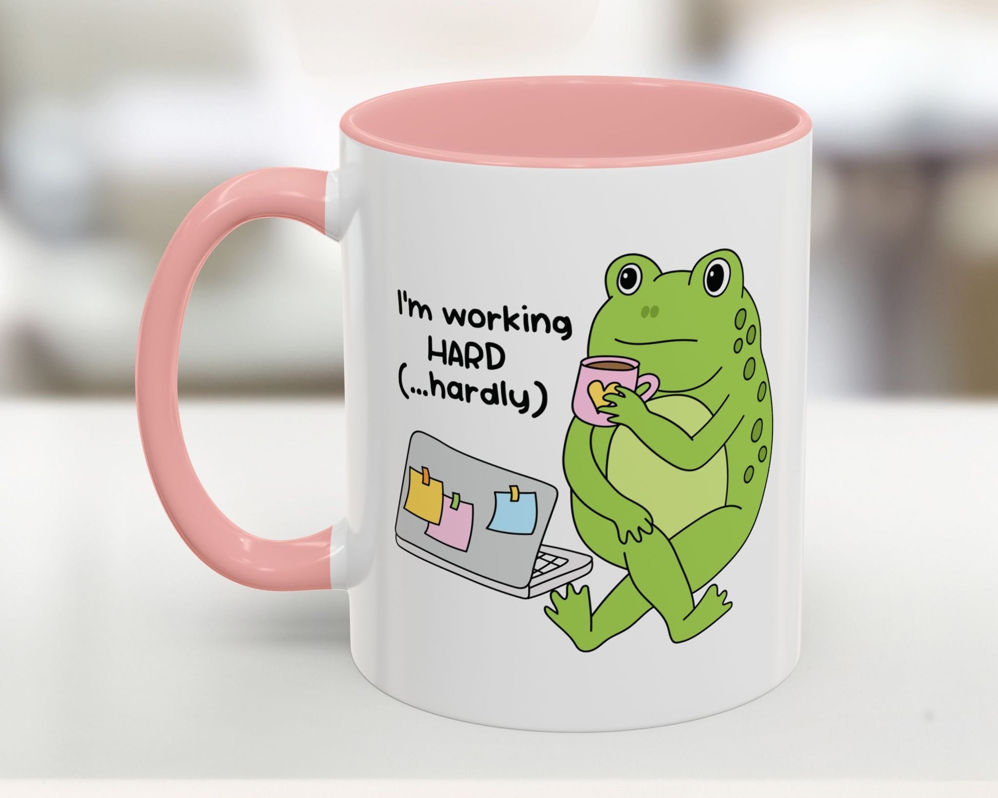 Mug with a green frog holding a cup and text 'I'm working HARD (...hardly)' on a white background.