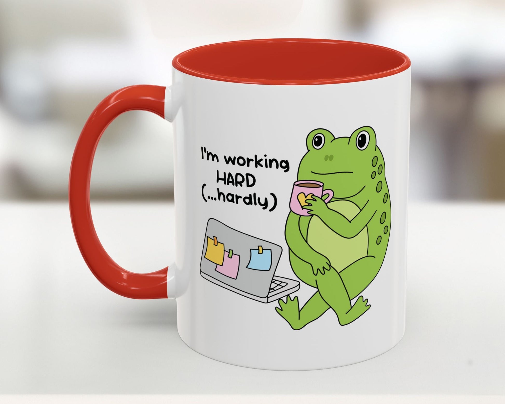 Mug with a green frog holding a cup and text 'I'm working HARD (...hardly)' on a white background.