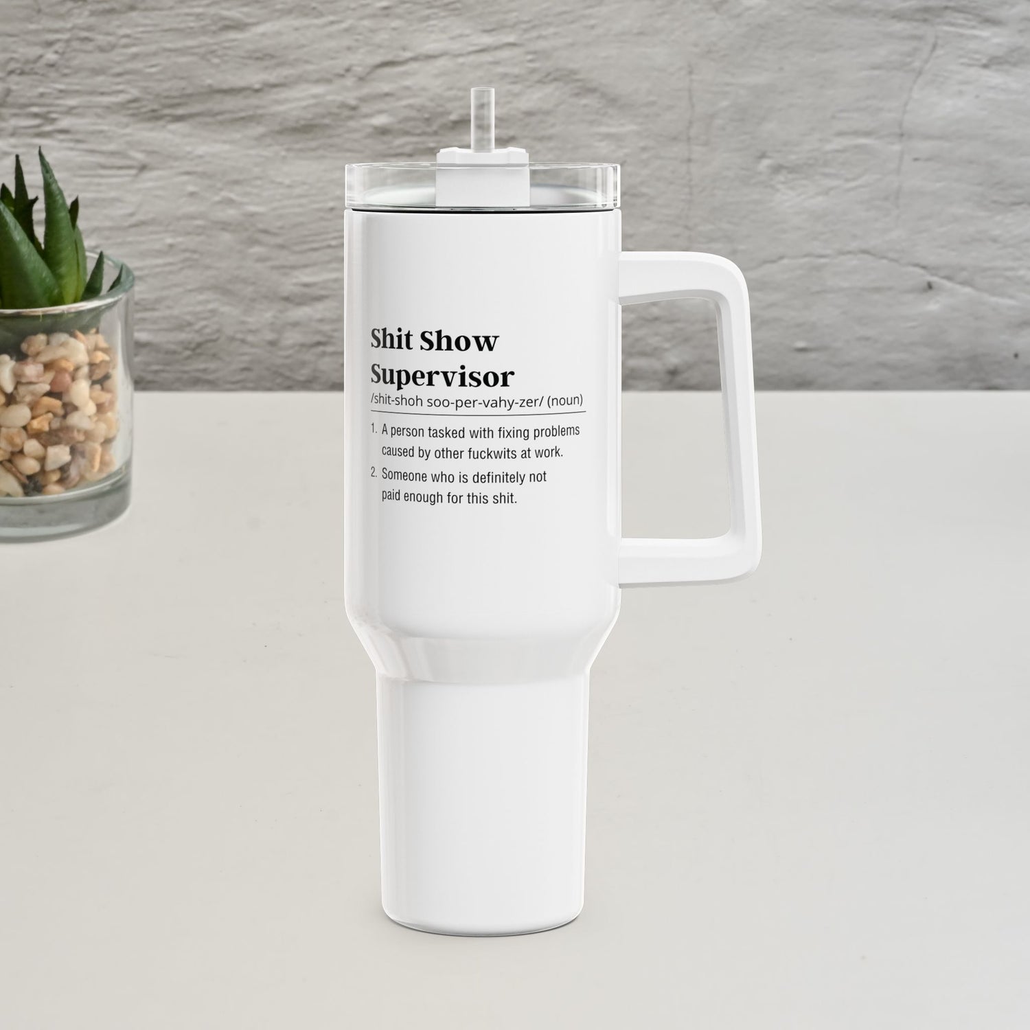 Shit show supervisor White tumbler with text on a gray surface with a plant in the background