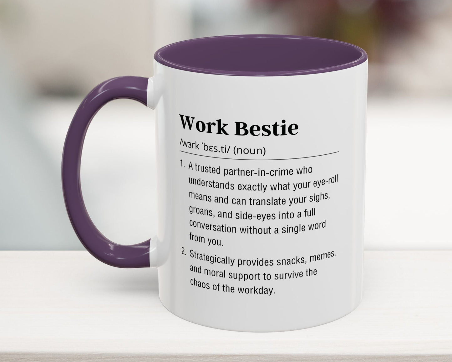 Mug with purple interior and handle displaying a definition of 'Work Bestie' on a blurred background.