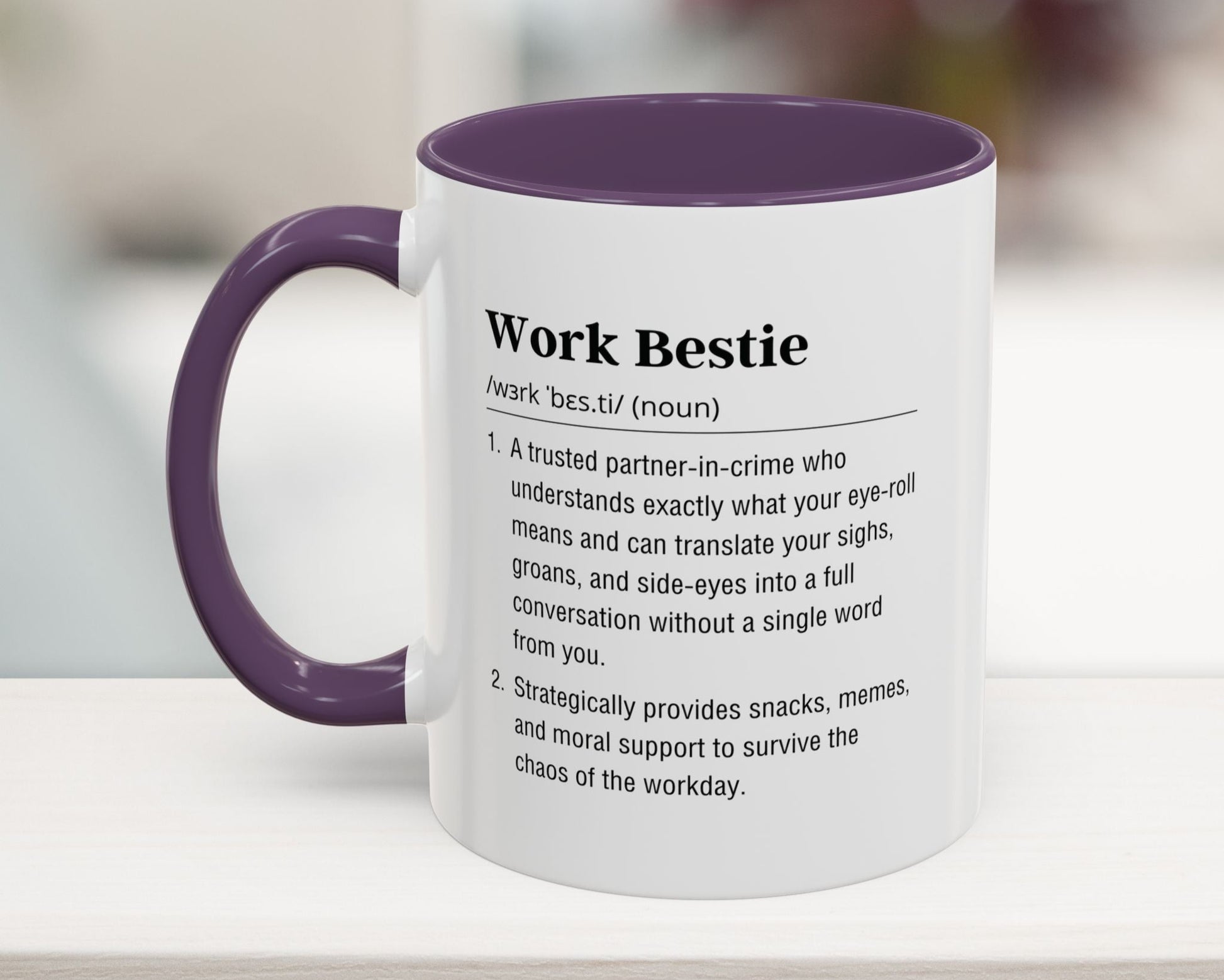Mug with purple interior and handle displaying a definition of 'Work Bestie' on a blurred background.
