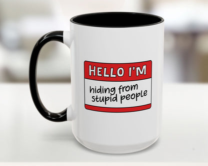 White mug with black interior and handle, featuring a red name tag with text on a blurred background.