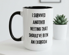 Mug with text 'I survived another meeting that should've been an exorcism' on a white surface with a plant in the background.