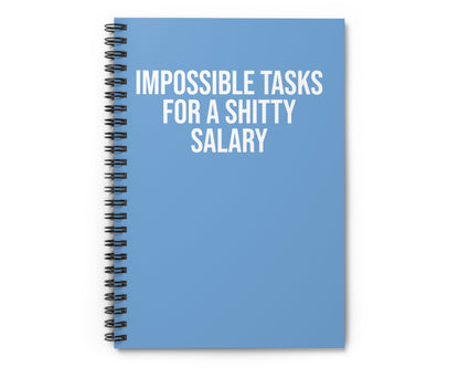 Blue notebook with text 'IMPOSSIBLE TASKS FOR A SHITTY SALARY' on a white background