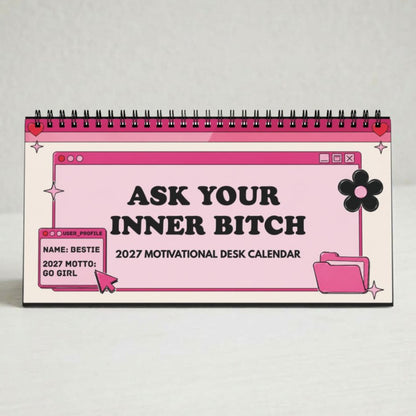 A pink and white spiral-bound desk calendar with black text reading 'ASK YOUR INNER BITCH 2027 MOTIVATIONAL DESK CALENDAR' and cartoon-style folder and flower graphics, photographed on a light neutral background.