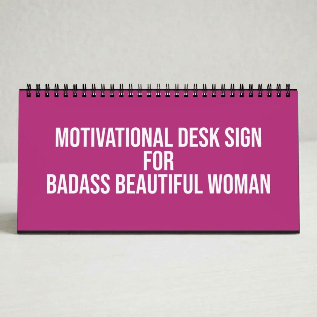 A pink spiral-bound desk sign with bold white text reading 'MOTIVATIONAL DESK SIGN FOR BADASS BEAUTIFUL WOMAN' on a light neutral background.