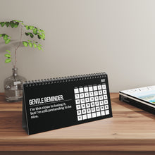 Desk calendar with humorous message on a wooden surface with a plant and magazine in the background.