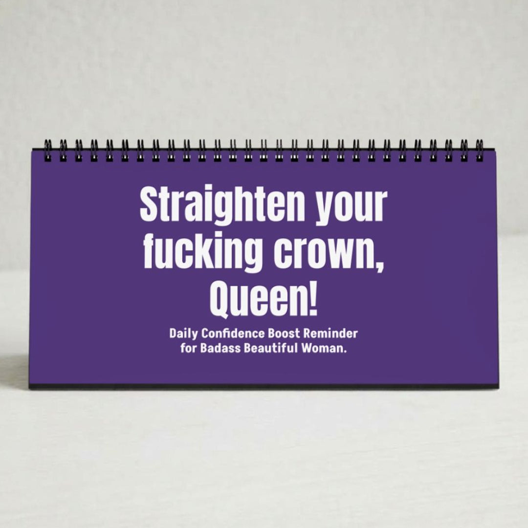 A purple spiral-bound desk calendar or planner standing upright on a light gray surface. The front cover has bold white text reading, 'Straighten your fucking crown, Queen!' with smaller text below, 'Daily Confidence Boost Reminder for Badass Beautiful Woman.