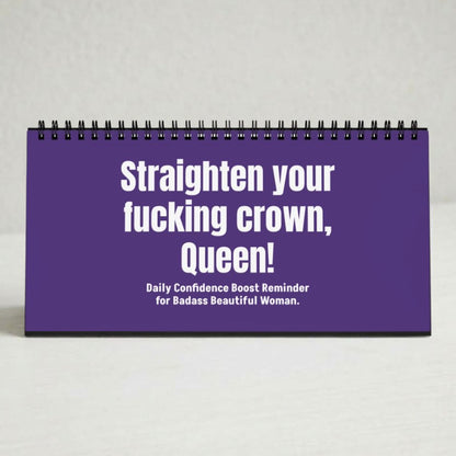 A purple spiral-bound desk calendar or planner standing upright on a light gray surface. The front cover has bold white text reading, 'Straighten your fucking crown, Queen!' with smaller text below, 'Daily Confidence Boost Reminder for Badass Beautiful Woman.