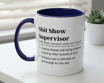 Mug with definition of 'Shit Show Supervisor' on a white surface with a plant in the background