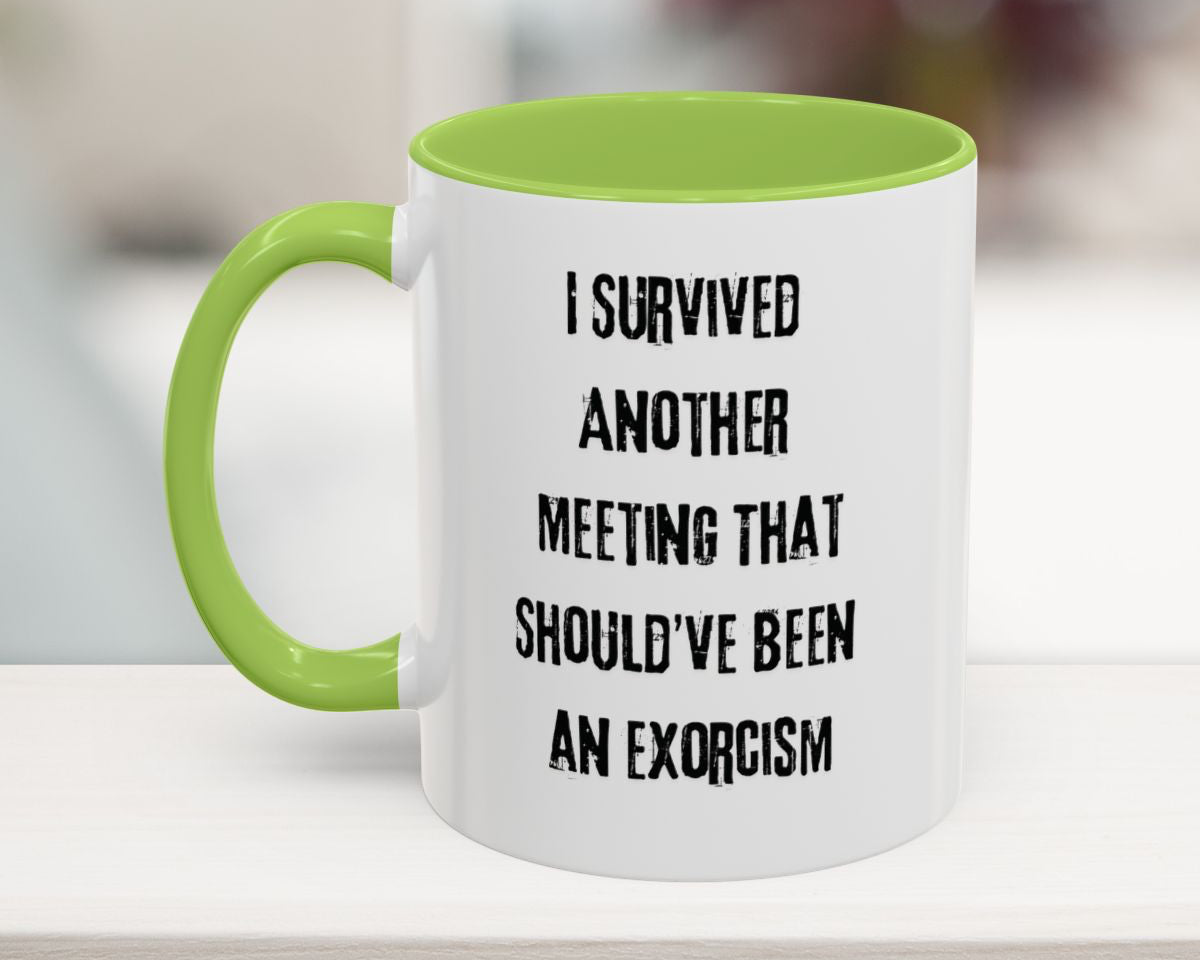 White mug with green handle and text 'I survived another meeting that should've been an exorcism' on a blurred background
