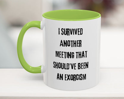 White mug with green handle and text 'I survived another meeting that should've been an exorcism' on a blurred background