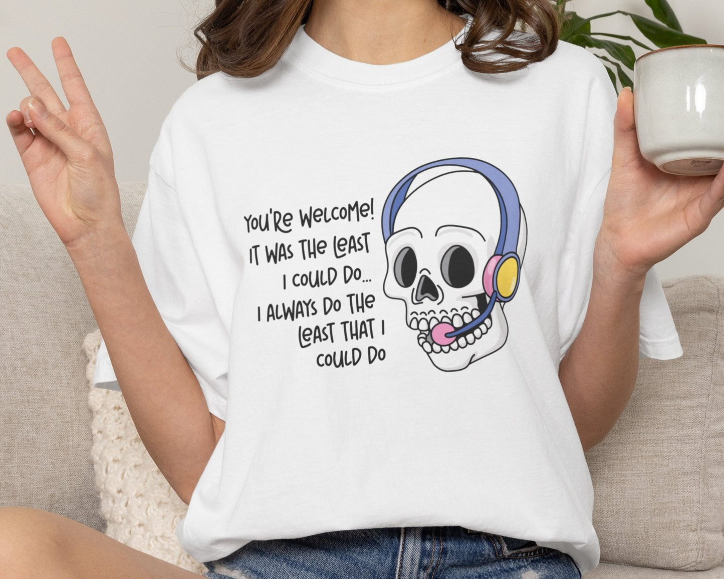 Person wearing a white t-shirt with a skull graphic and text, holding a mug.