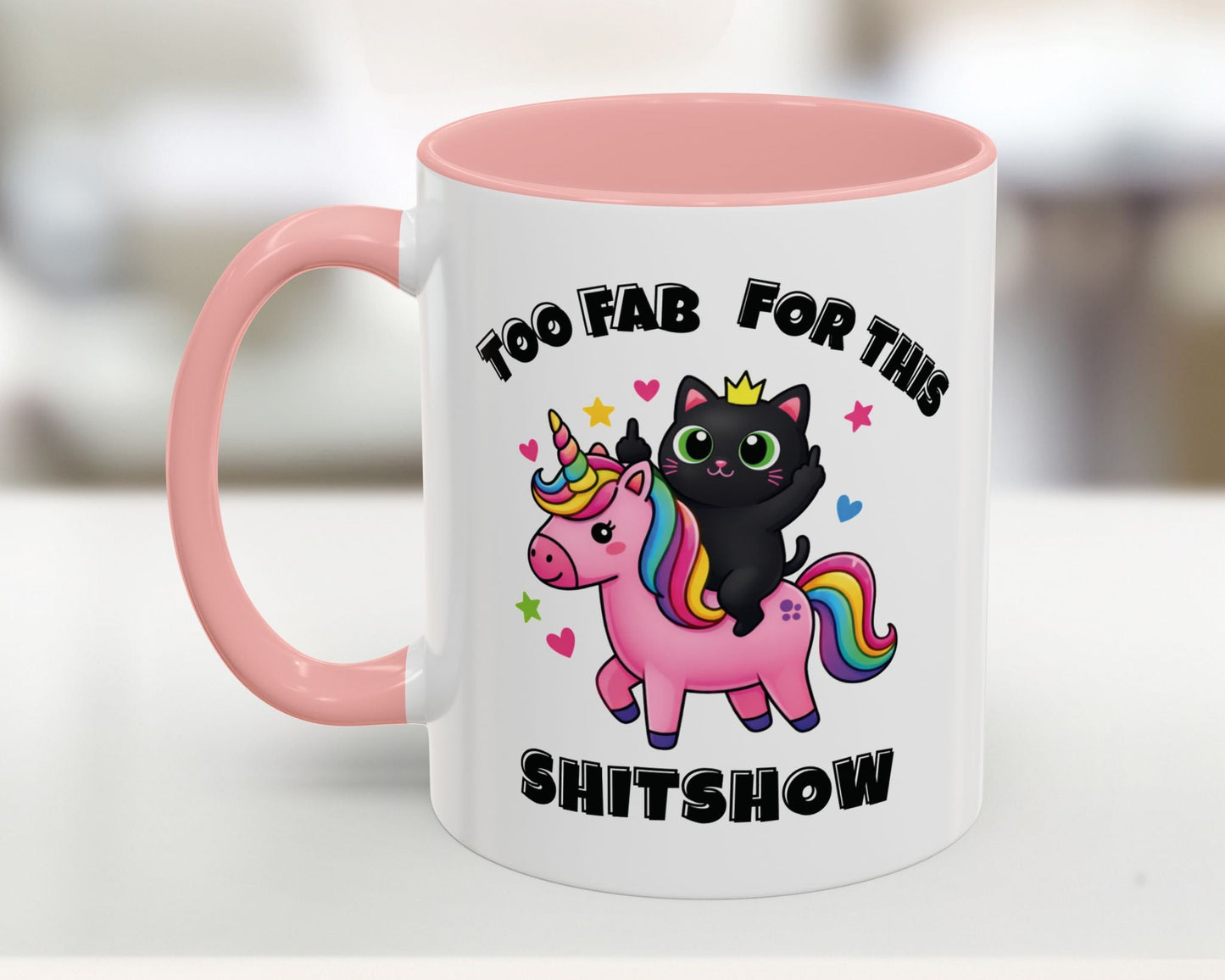Mug with a black cat on a unicorn, pink handle, and text 'Too Fab for this Shitshow' on a blurred background.