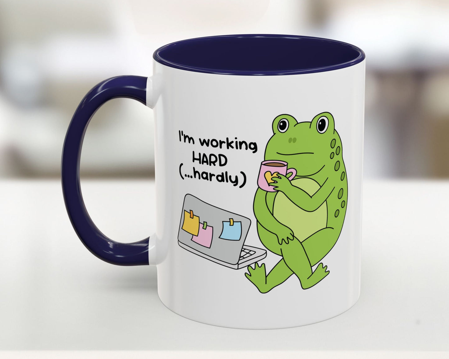 Mug with a green frog holding a cup and text 'I'm working HARD (...hardly)' on a white background.