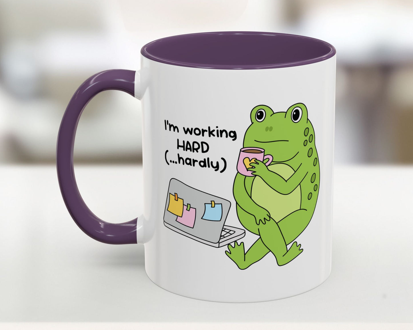 Mug with a green frog holding a cup and text 'I'm working HARD (...hardly)' on a white background.
